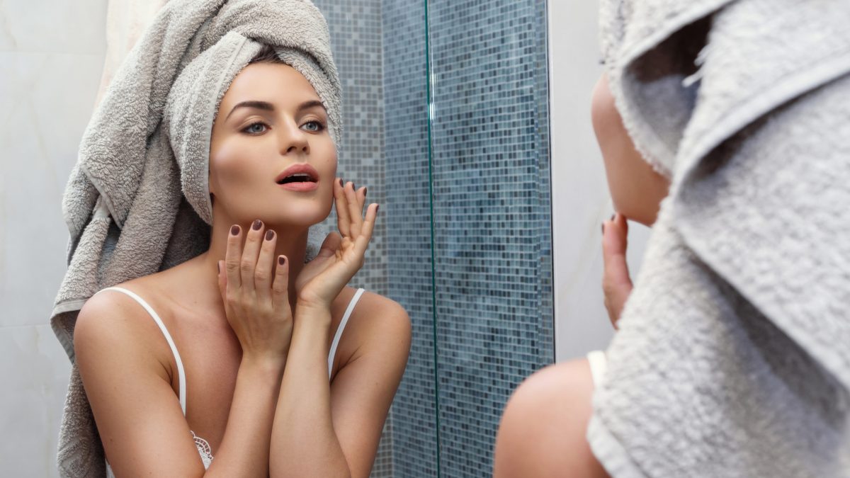 Beautiful woman with a towel on her head, looking in the mirror and applying make up