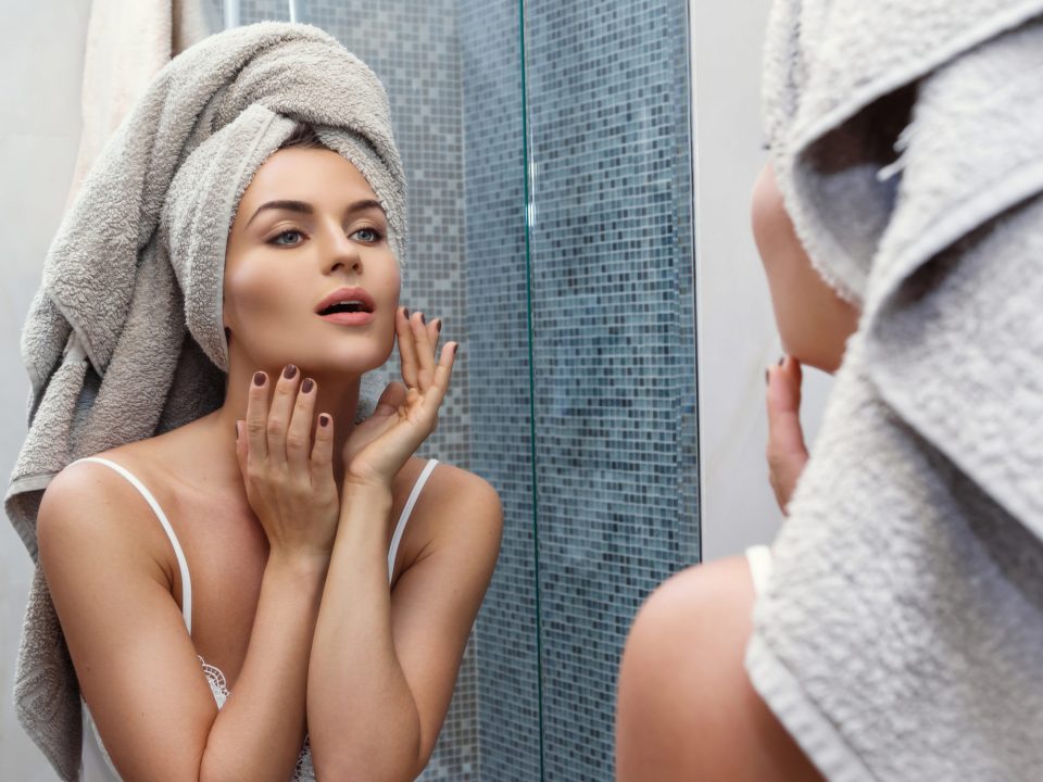 Beautiful woman with a towel on her head, looking in the mirror and applying make up